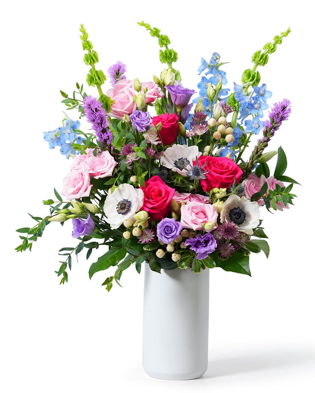 A vibrant floral arrangement featuring pink and purple roses, anemones, and lush greenery in a sleek white vase.