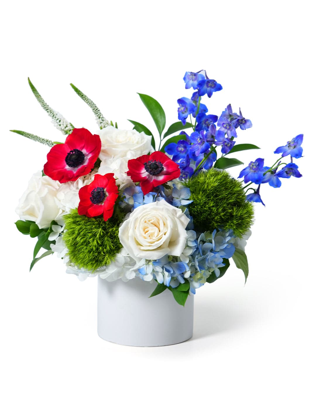 Vibrant floral arrangement featuring red anemones, blue delphiniums, and lush green accents in a sleek white vase.