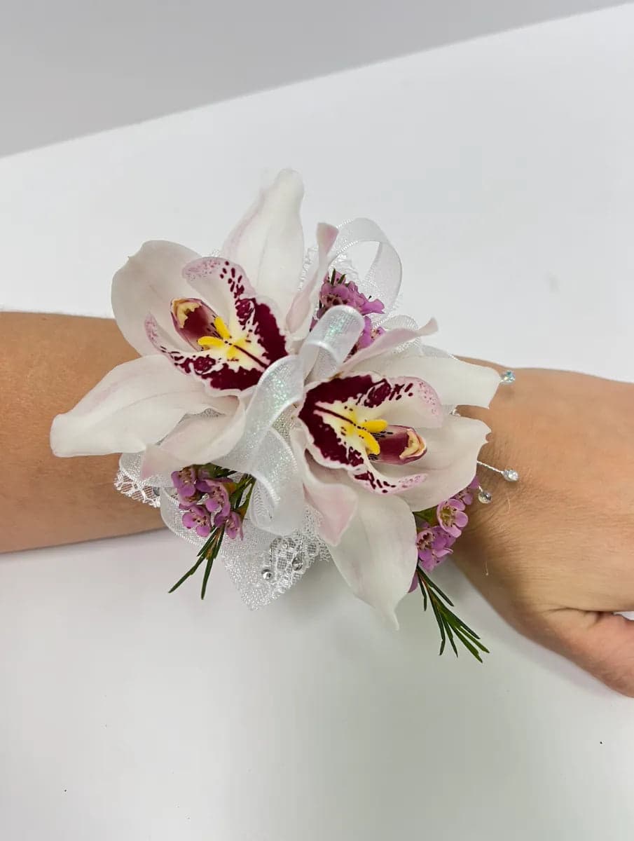 Elegant wrist corsage featuring two white orchids and delicate lavender flowers, adorned with lace and a ribbon.